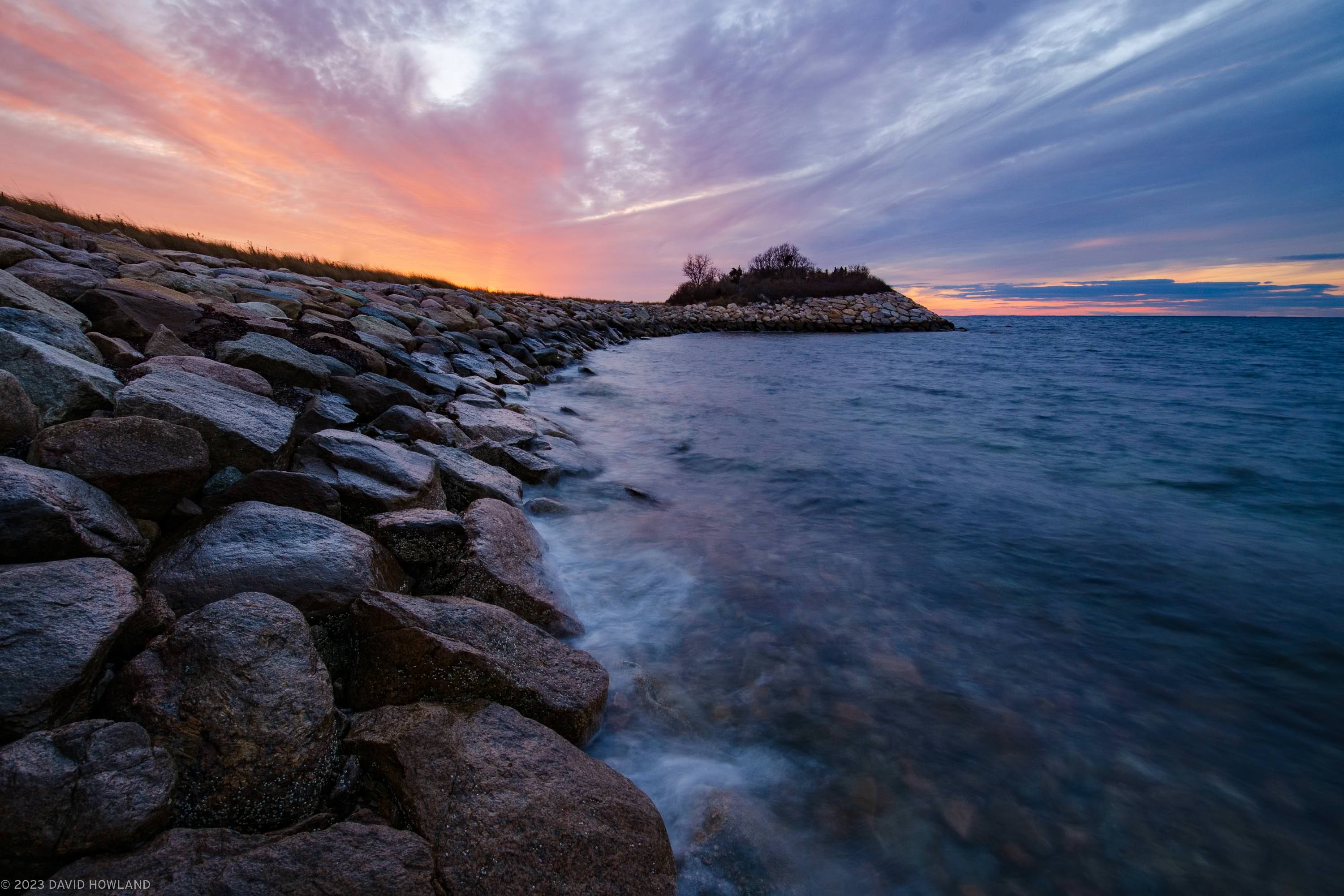 A photo of a colorful sunset behind the rocky outcropping of the Knob in Woods Hole, Massachusetts on Cape Cod.