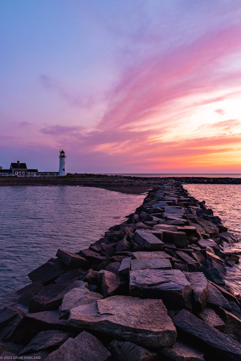 A photo of a rock jetty and Scituate Lighthouse in front of a colorful sunrise.