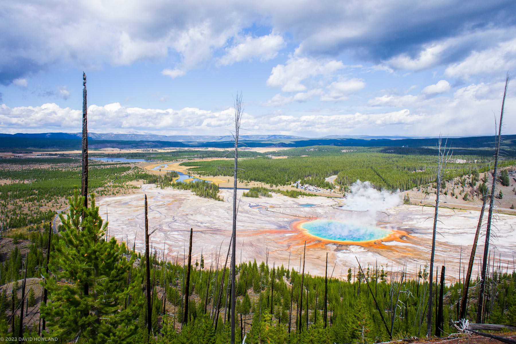 A photo of the colorful hot springs of Grand Prismatic Sping in Yellowstone National Park seen from above on a nearby ridgeline.