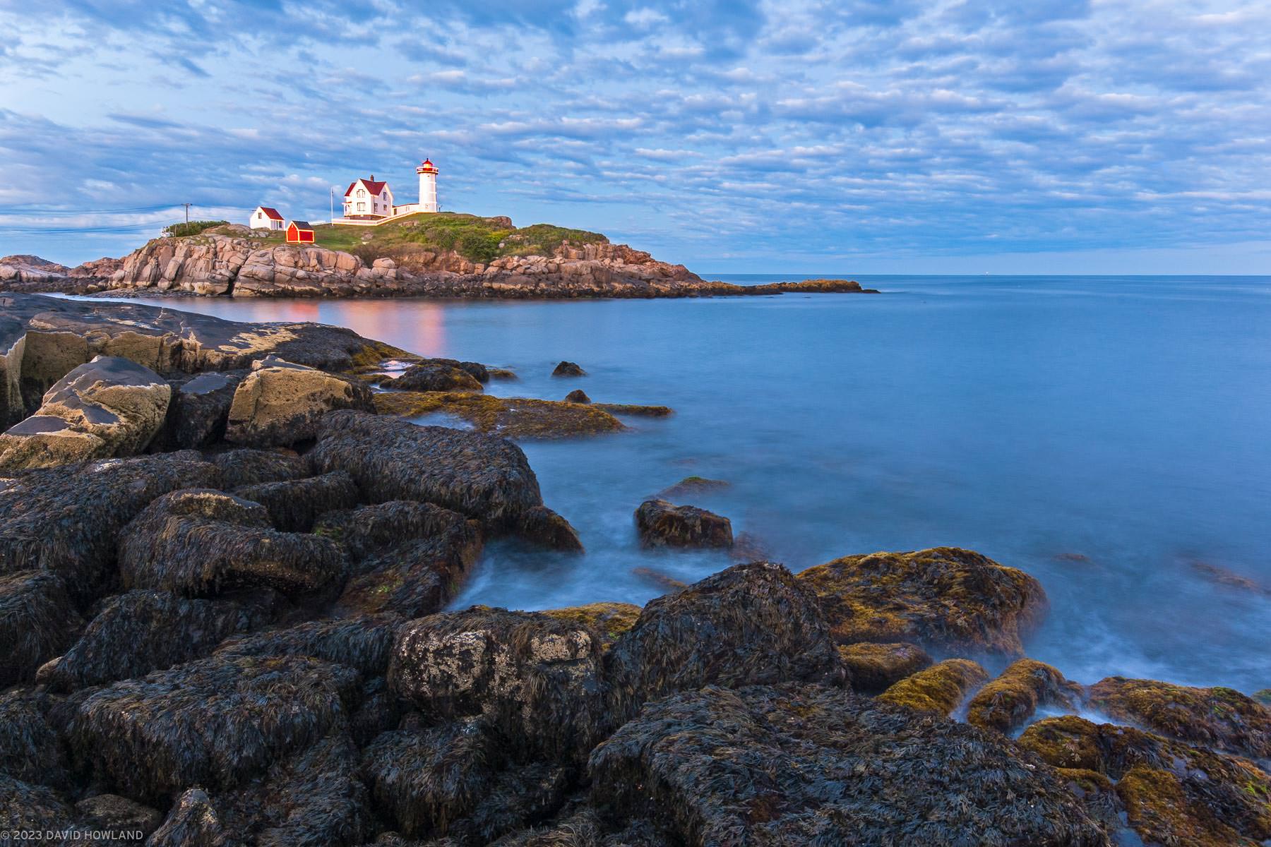 A photo of seaweed covered rocks on the coast of Maine with Nubble Lighthouse in the background.