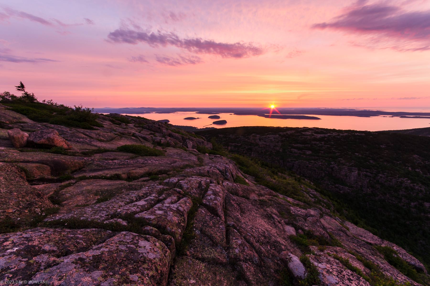 A photo of the sun rising over the calm waters of Frenchman Bay and lighthing up the cracked granite slopes of Cadillac Mountain in Acadia National Park in Maine.