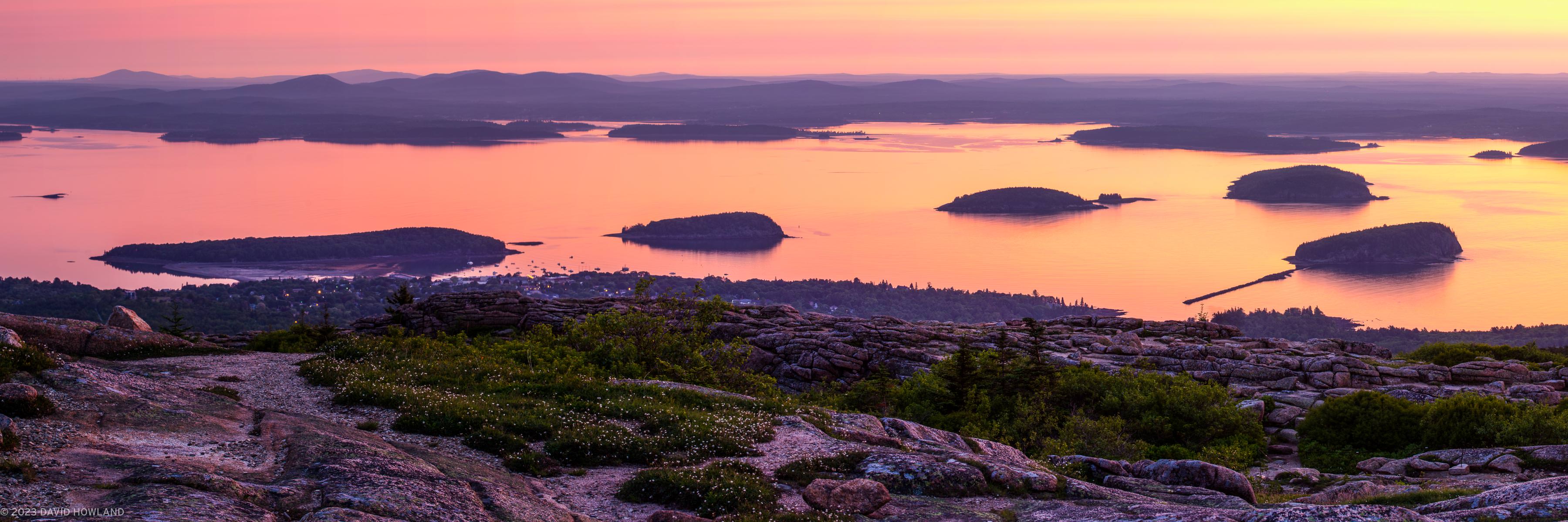 A panorama photo of the sun rising over the calm waters of Frenchman Bay, the coastal town of Bar Harbor, and the wildflowers at the granite summit of Cadillac Mountain in Acadia National Park