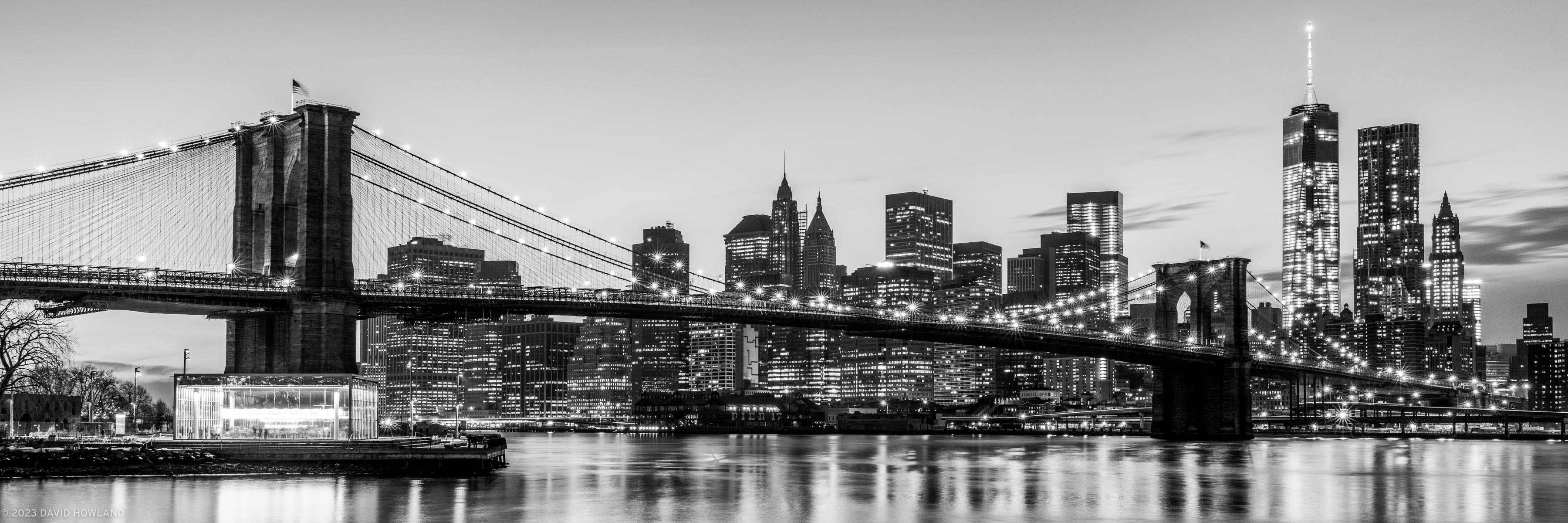 A black and white panorama photo of the Brooklyn Bridge in front of the illuminated skyscrapers of downtown New York City at night.