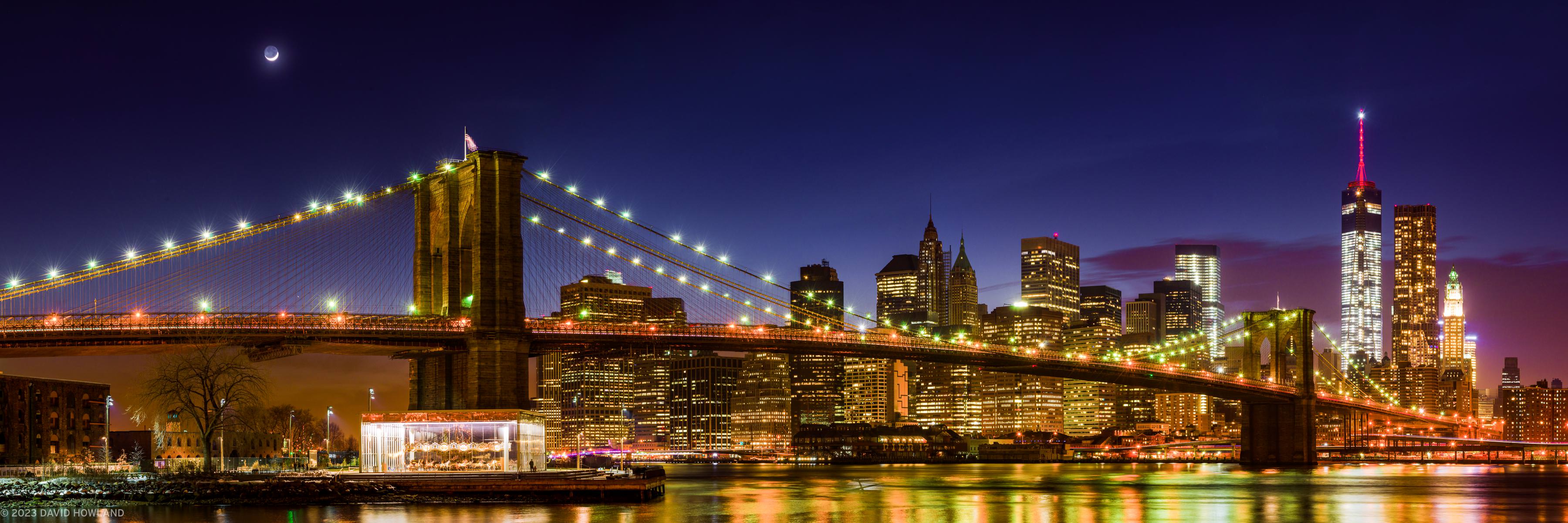 A panorama photo of a crescent moon rising over the Brooklyn Bridge and illuminated skyscrapers of downtown New Yok City at night.