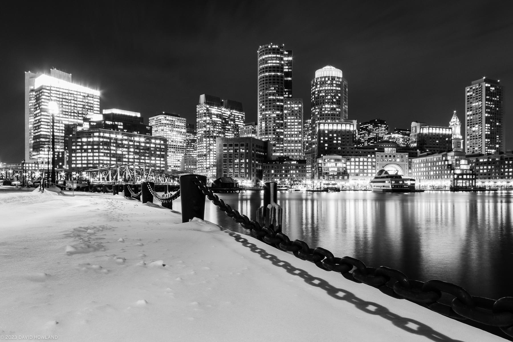 A black and white photo of the illuminated Boston skyline above the waters of Boston Harbor and a snow covered walkway.