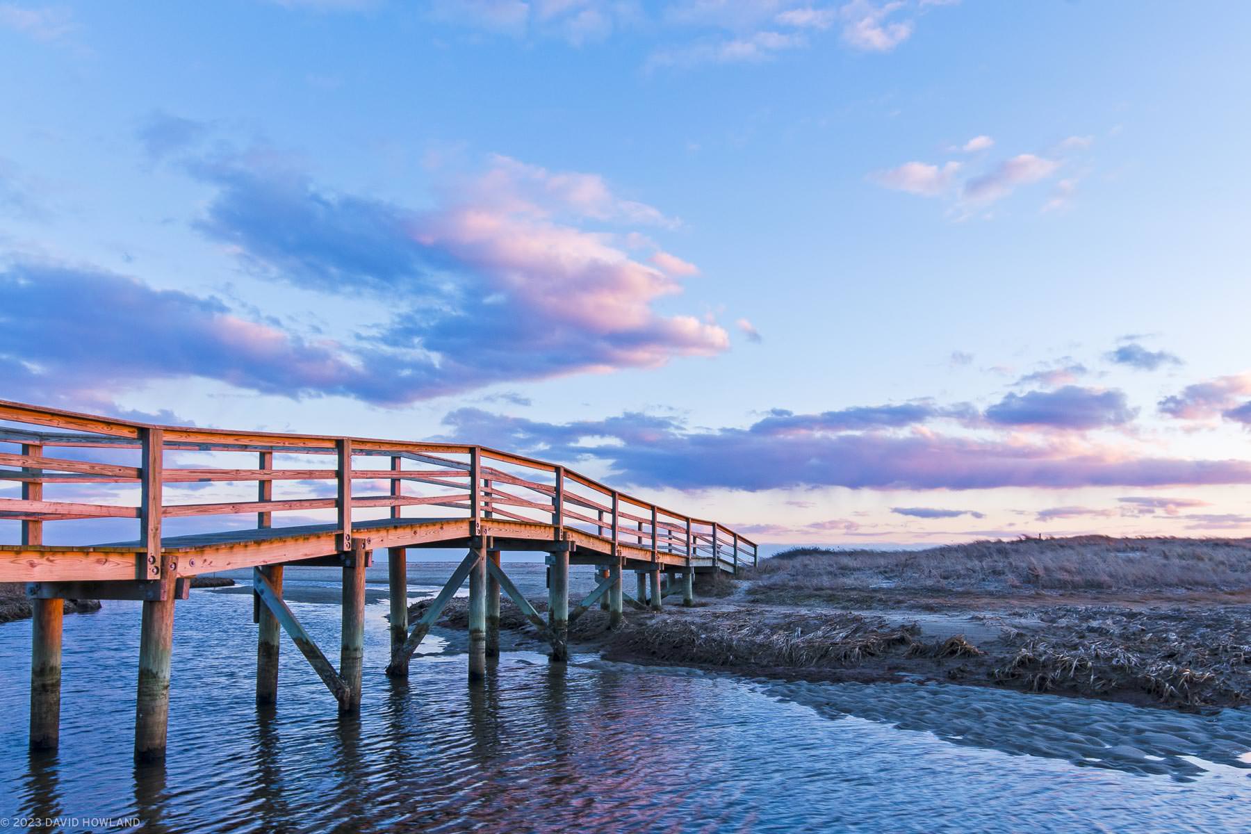 A photo of sunset lighting up the clouds in the sky over a boardwalk crossing a tidal channel at Ridgevale Beach in Chatham, MA.