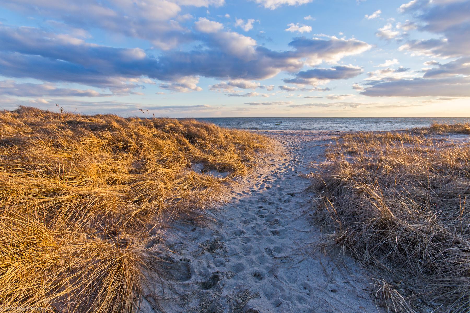 A photo of clouds in the sky over a dune grass covered beach at sunset.