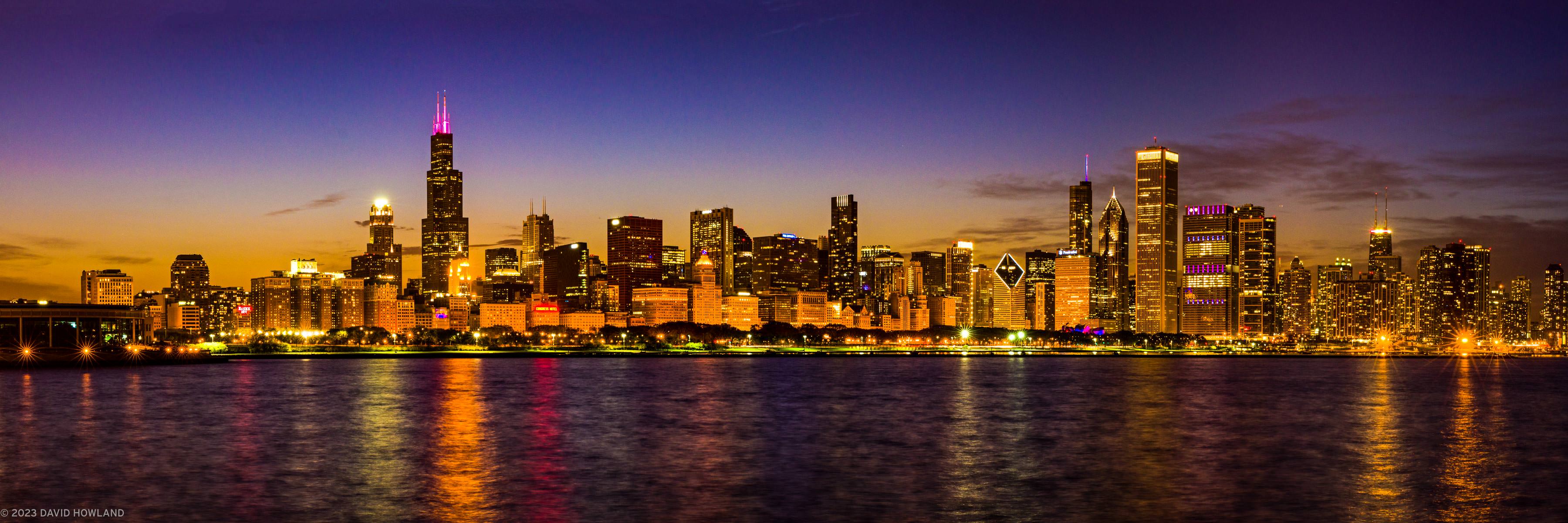 A panorama photo of the skyscrapers of the Chicago city skyline lit up against the fading orange sunset light.