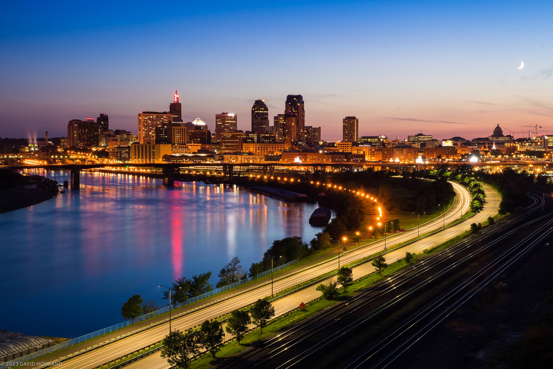 A photo of the skyline of Saint Paul, Minnesota at sunset with the Mississippi River, a road, and railroad tracks curving through the foreground