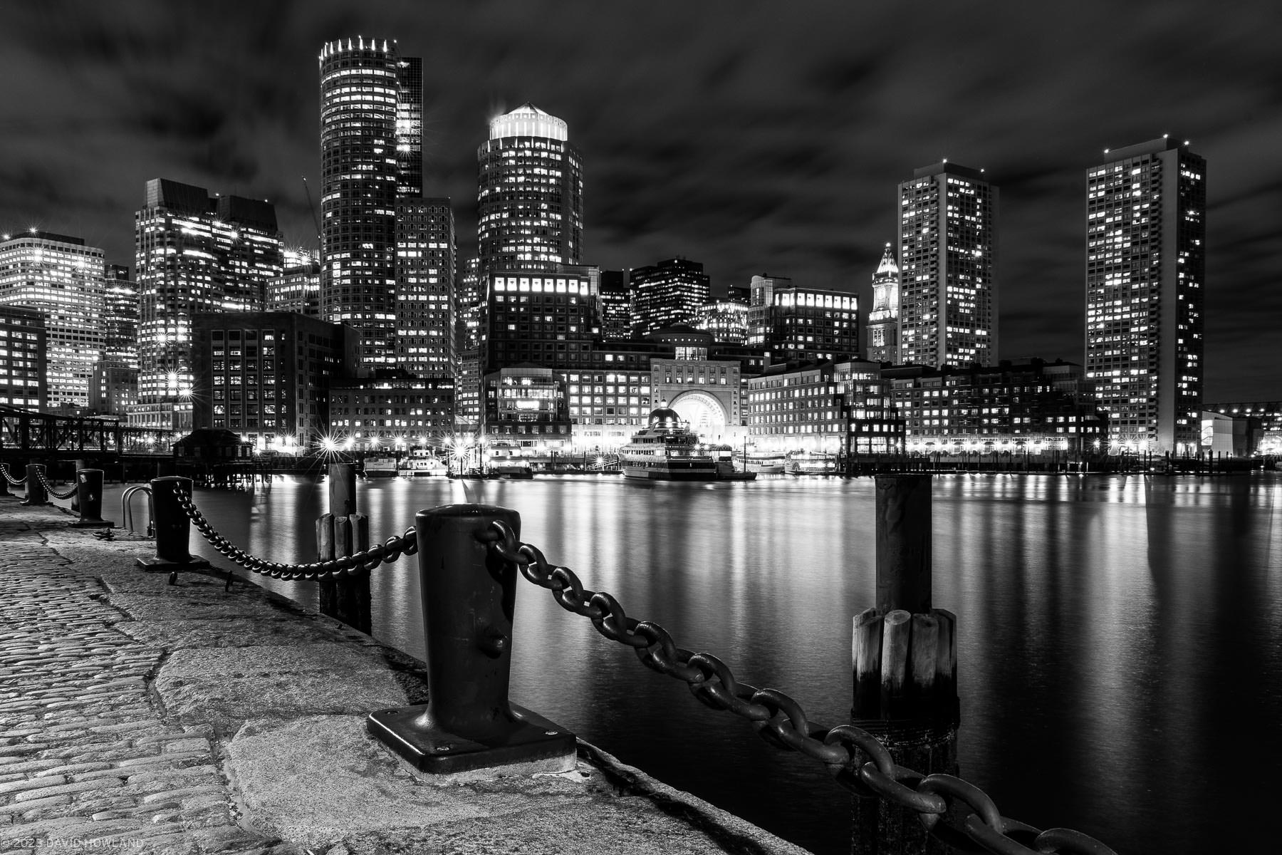 A black and white photo of the downtown Boston skyline with boats and Boston Harbor in the foreground.