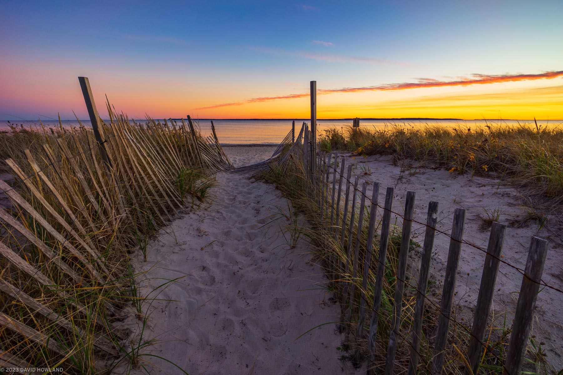 A photo of a battered wooden fence marking a path through the dunes of Surf Drive Beach at sunset in Falmouth, Massachusetts.