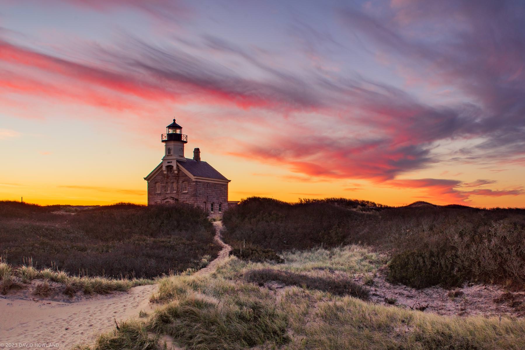 The clouds behind North Lighthouse on Block Island glow red and orange at sunrise.