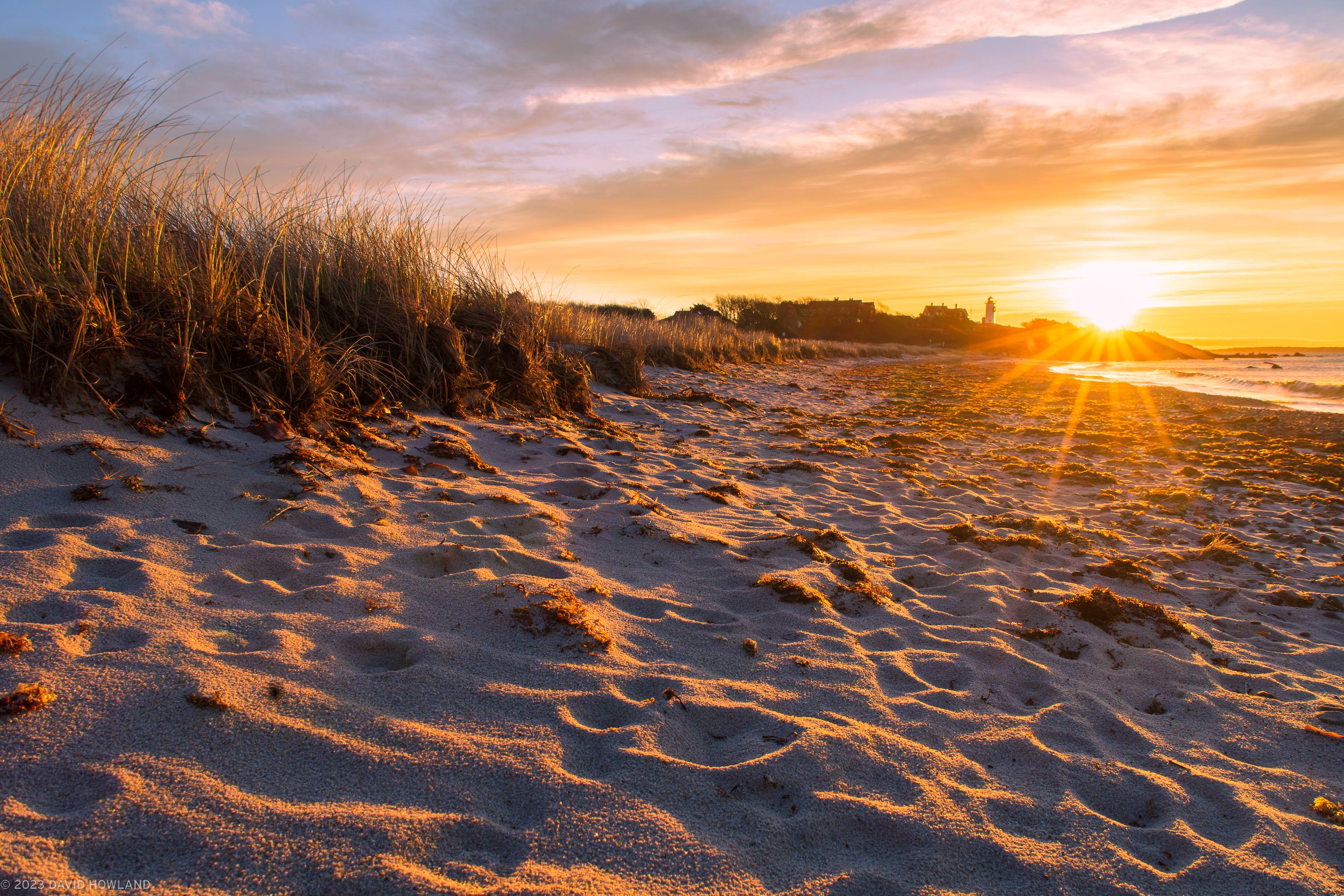 A photo of the sun rising behind Nobska Lighthouse and the sand and beach grass of Nobska Beach in Woods Hole, Massachusetts on Cape Cod.