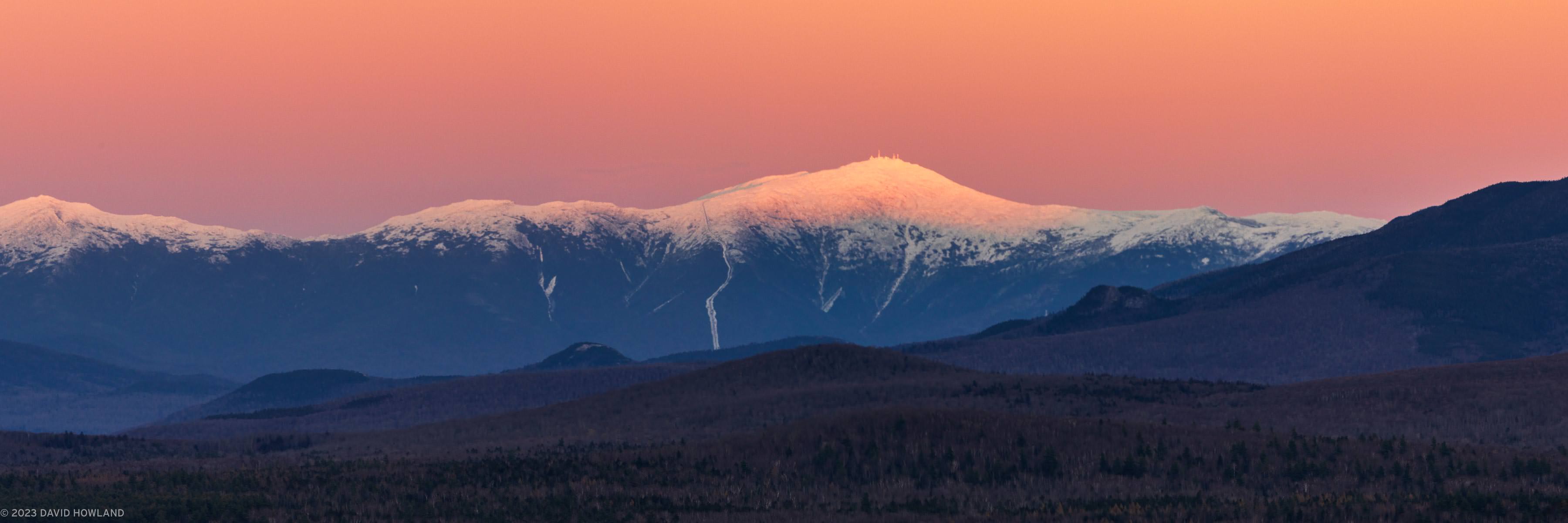 A panorama photo of sunset making the snow-covered Presidential mountain range in the White Mountains of New Hampshire glow orange.
