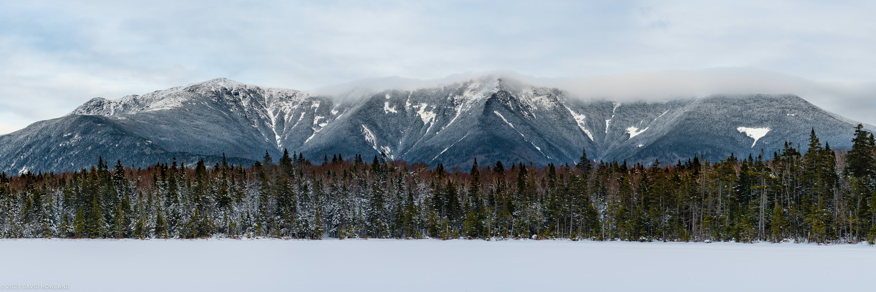 A panorama photo of the snow covered Franconia Ridge mountains above a pine forest and frozen Lonesome Lake in Franconia Notch, New Hampshire.