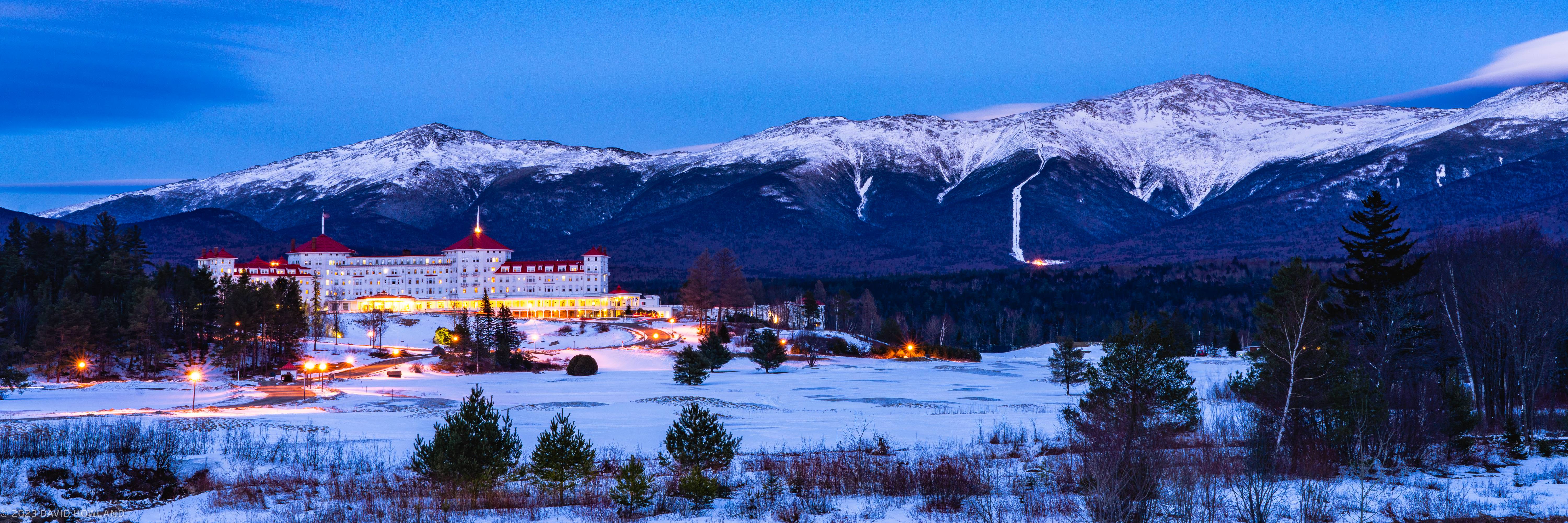 A panorama photo of the glowing orange lights of the Mount Washington Hotel building in front of the snow covered Presidential mountain range in New Hampshire.
