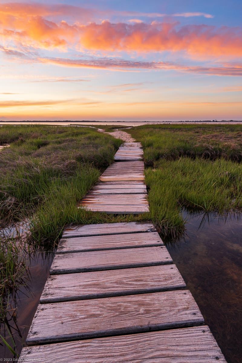 A photo of a boardwalk in a salt marsh under glowing orange skies at sunset in Barnstable, Massachusetts on Cape Cod.