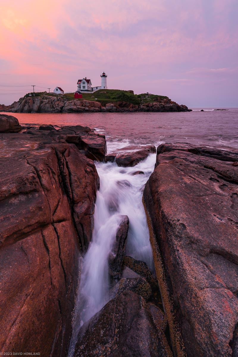 A sunset photo of waves crashing on the rocks of the coast of Maine with Nubble Lighthouse in the background.