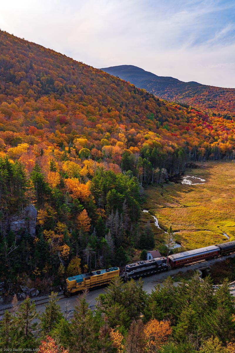 A photo of a train pulled by two diesel engines in front of the colorful yellow and red foliage on the trees of the White Mountains in New Hampshire.