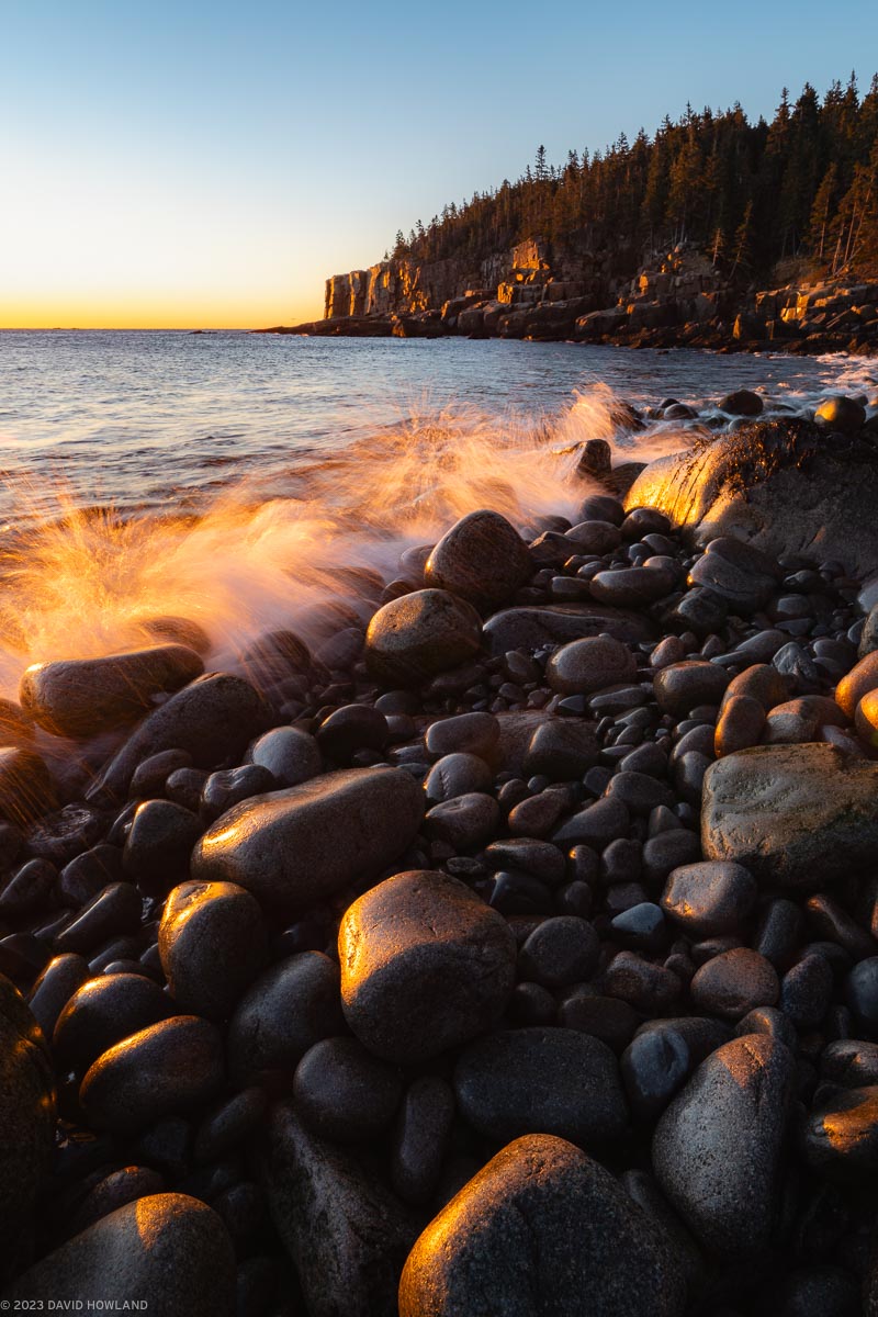 A photo of a waves breaking onto the smooth boulders and granite cliffs of the coastline of Acadia National Park in Maine.