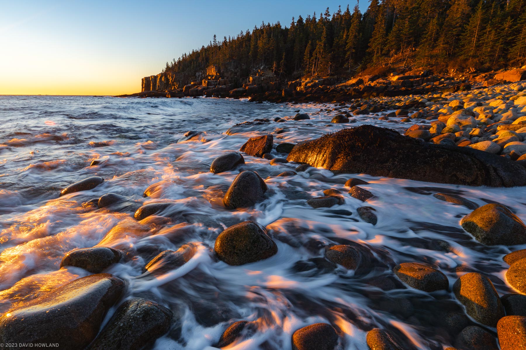 A photo of a waves crashing onto the smooth rocks and granite cliffs of the coastline of Acadia National Park in Maine.