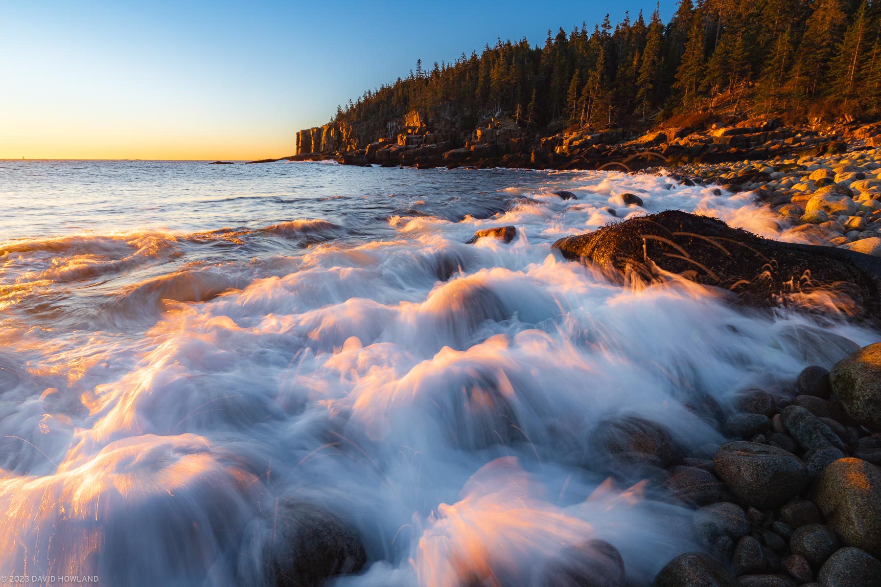 A photo of a waves breaking onto the smooth boulders and granite cliffs of the coastline of Acadia National Park in Maine.