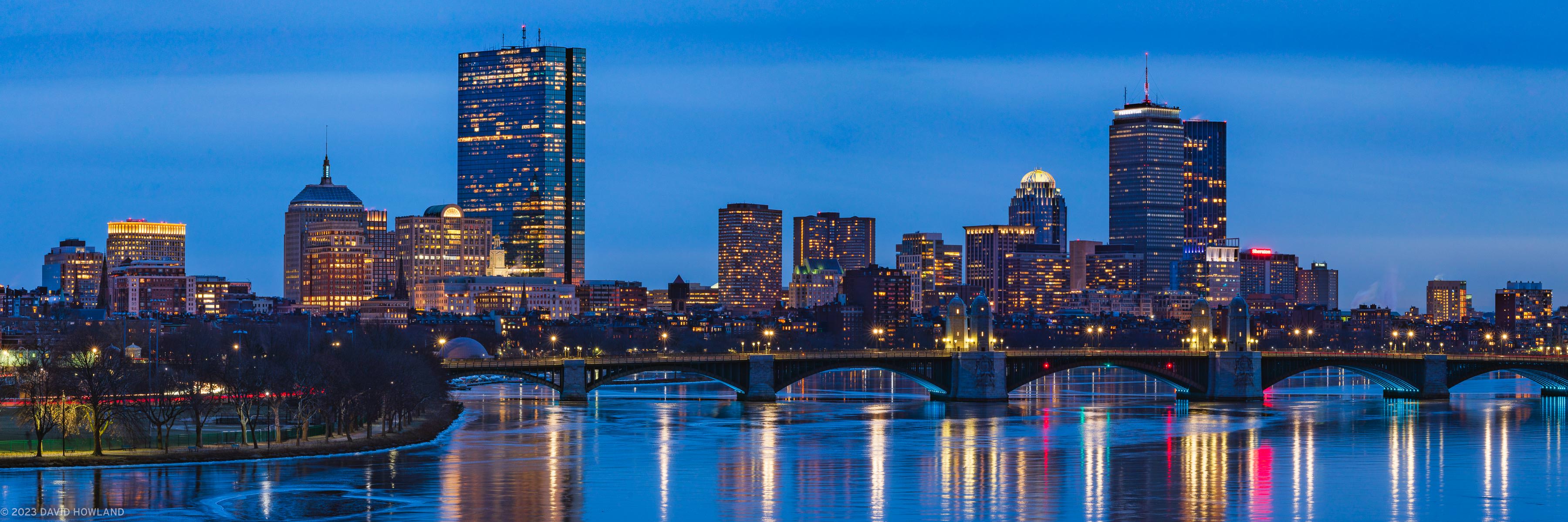 A panorama photo of the city lights of the Boston skyline and the Longfellow Bridge spanning a frozen Charles River at dusk