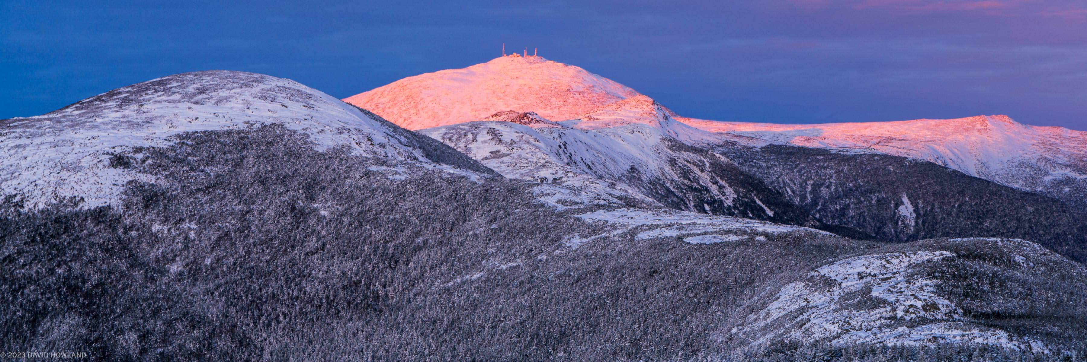 A panorama photo of orange alpenglow lighting up Mount Washington and the other peaks of the Presidential Range in the White Mountains of New Hampshire.