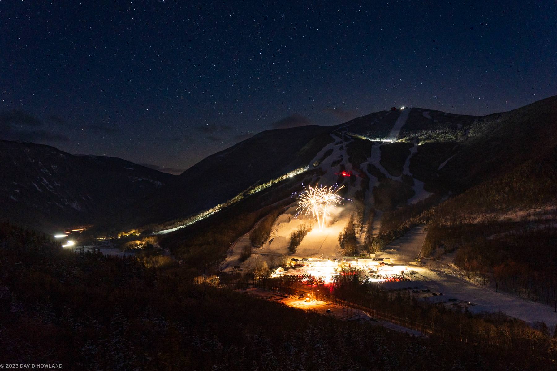 A photo of fireworks exploding over snow-covered Cannon Mountain under a clear winter sky filled with stars