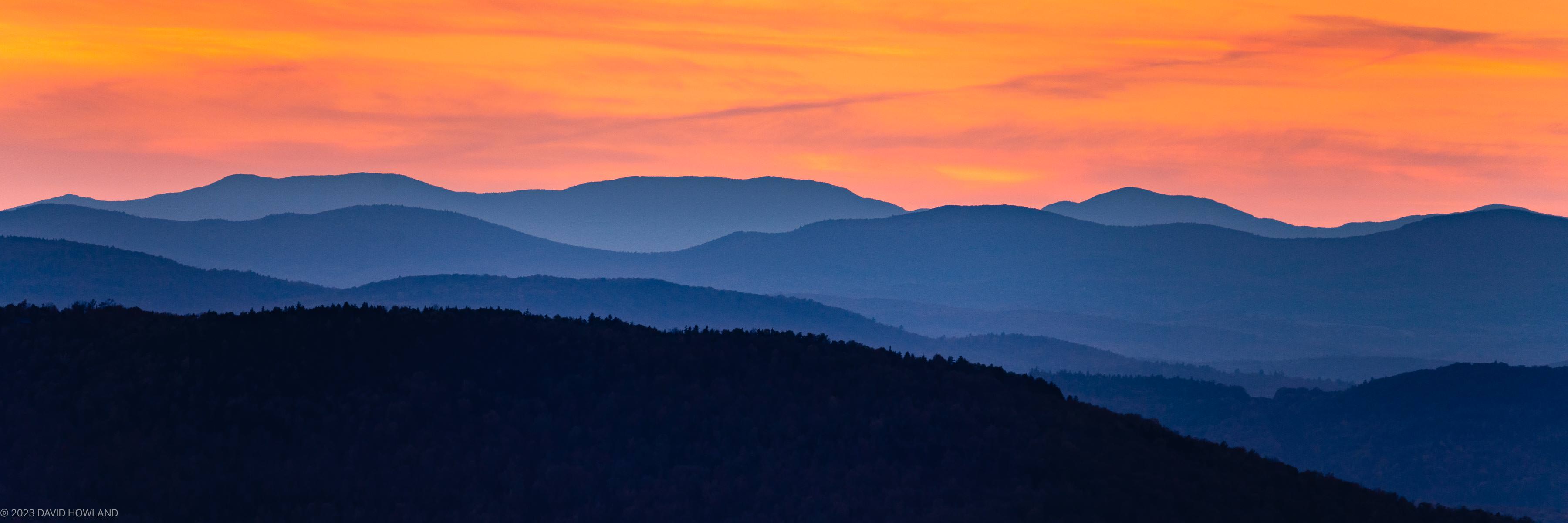 A colorful panorama photo of an orange sunset behind the layered blue-hued ridges of the Green Mountains of Vermont.