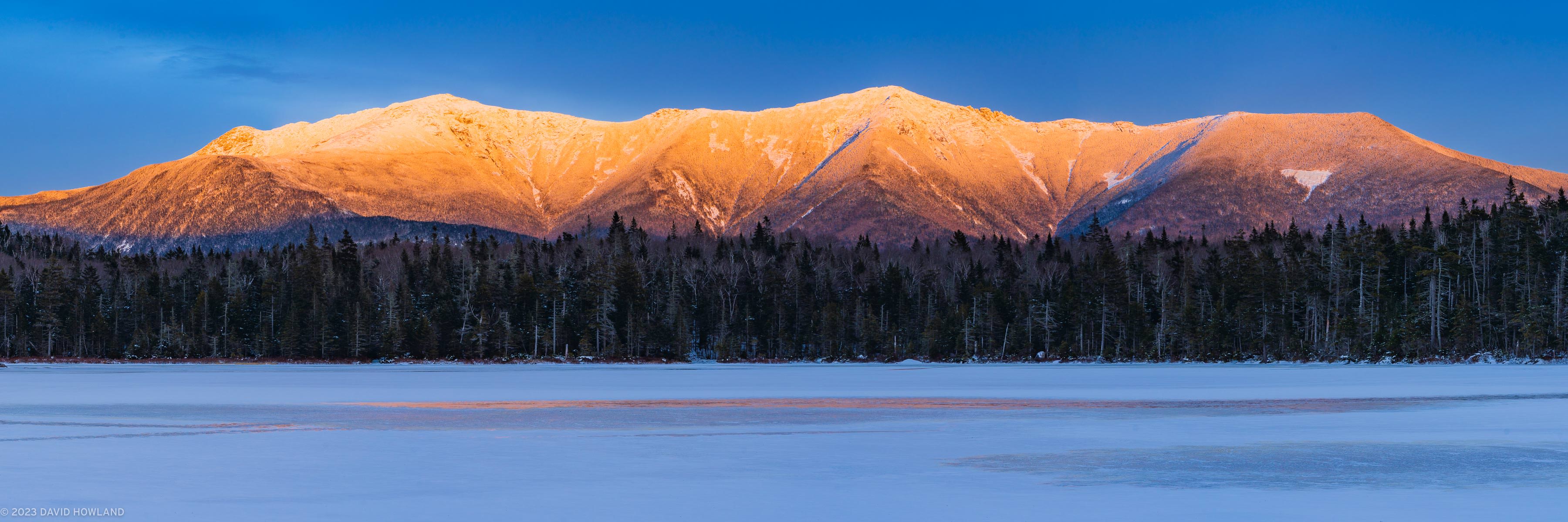 A panorama photo of the snow-covered peaks of Franconia Ridge glowing orange with alpenglow over the frozen surface of Lonesome Lake