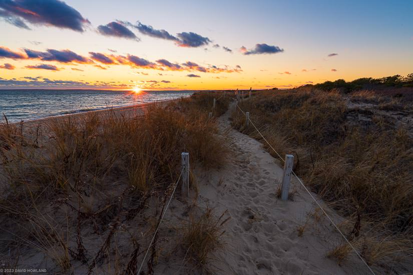 A photo of the sun setting behind the Atlantic Ocean and a sandy path through the dune grass of South Cape Beach State Park on Cape Cod.