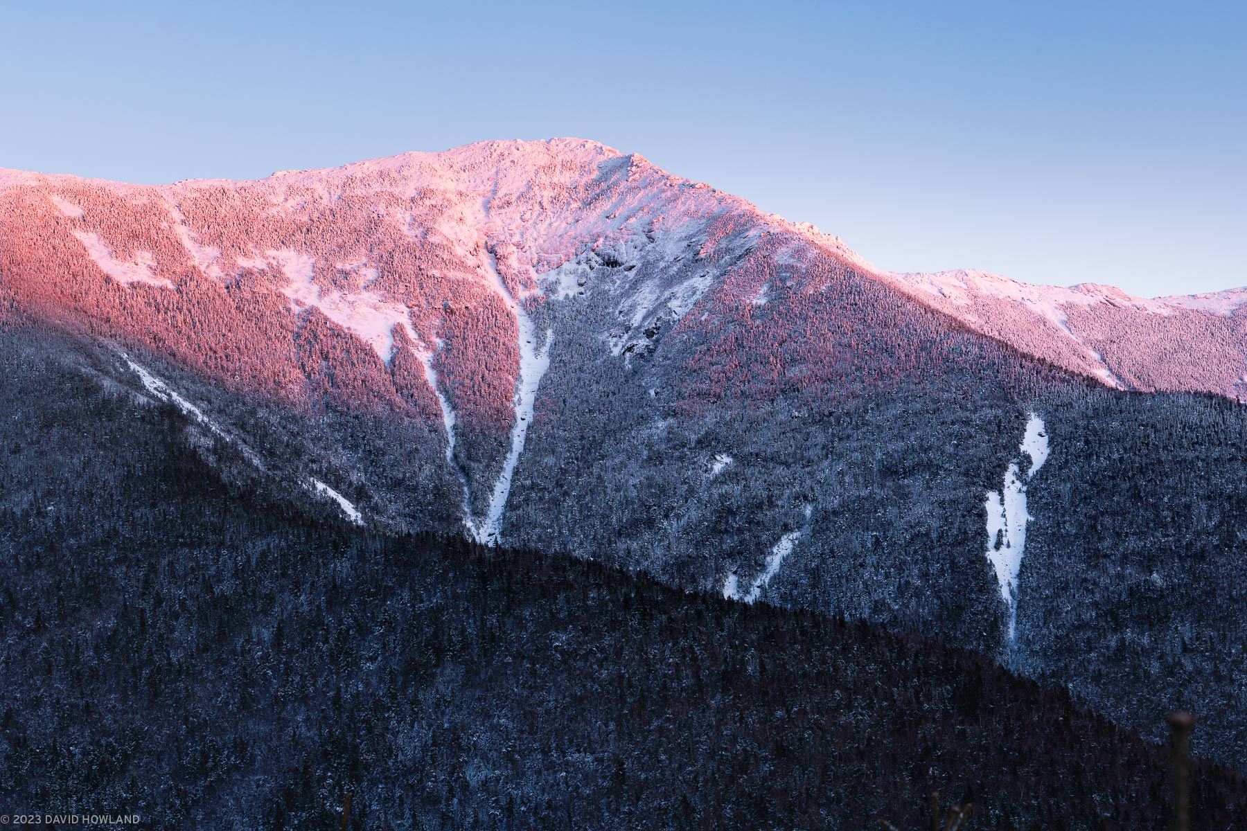 This winter landscape captures Mount Lincoln in New Hampshire's Franconia Notch during alpenglow - the rosy pink light that illuminates mountain peaks at sunset. The peak rises dramatically against a clear blue sky, with its summit bathed in pink-orange light while the lower slopes remain in cool blue shadow. Snow covers the mountainside, with several distinctive landslide paths visible as white strips cutting through the dark evergreen forest. The contrast between the warm-lit upper peak and the shadowed lower slopes creates a striking visual divide that emphasizes the mountain's height and grandeur.