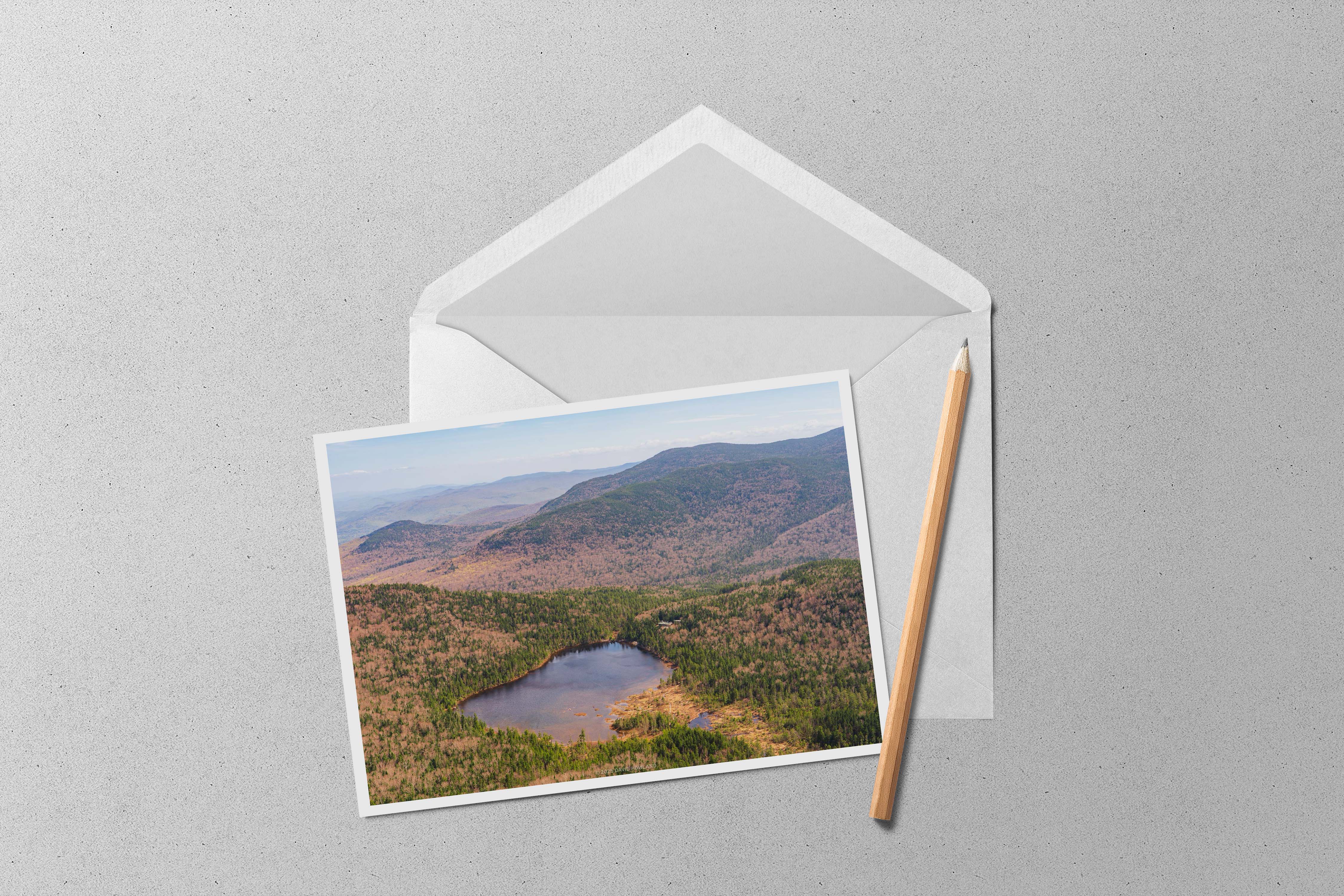 This landscape photograph captures a pristine mountain lake nestled within New England's rolling hills during spring, with the forested landscape displaying a mix of emerging green foliage and bare deciduous trees creating a patchwork of colors across the undulating terrain. The circular lake sits like a dark mirror in the valley below, surrounded by multiple ridgelines that fade into distant blue-gray mountain silhouettes under soft, diffused lighting.