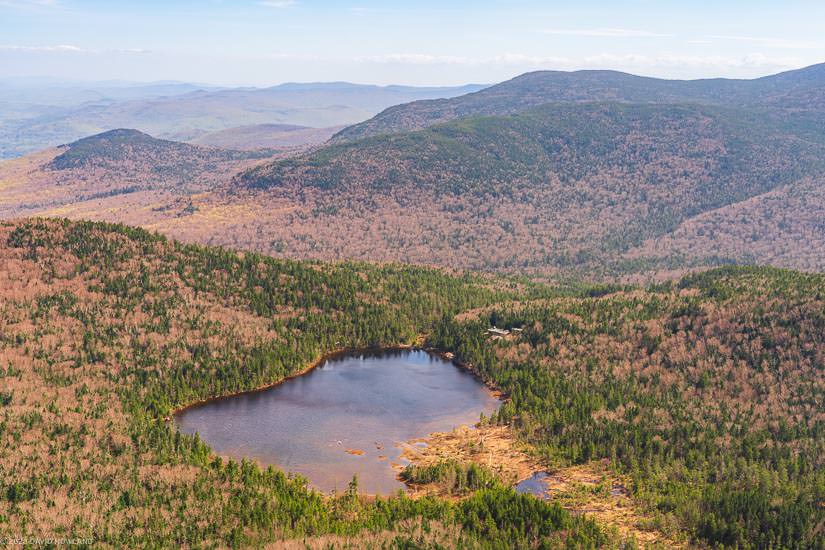 This landscape photograph captures a pristine mountain lake nestled within New England's rolling hills during spring, with the forested landscape displaying a mix of emerging green foliage and bare deciduous trees creating a patchwork of colors across the undulating terrain. The circular lake sits like a dark mirror in the valley below, surrounded by multiple ridgelines that fade into distant blue-gray mountain silhouettes under soft, diffused lighting.