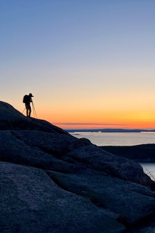 David Howland silhouetted behind a camera and tripod on a granite summit above the ocean at twilight.