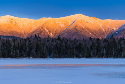 A panorama photo of the snow-covered peaks of Franconia Ridge glowing orange with alpenglow over the frozen surface of Lonesome Lake