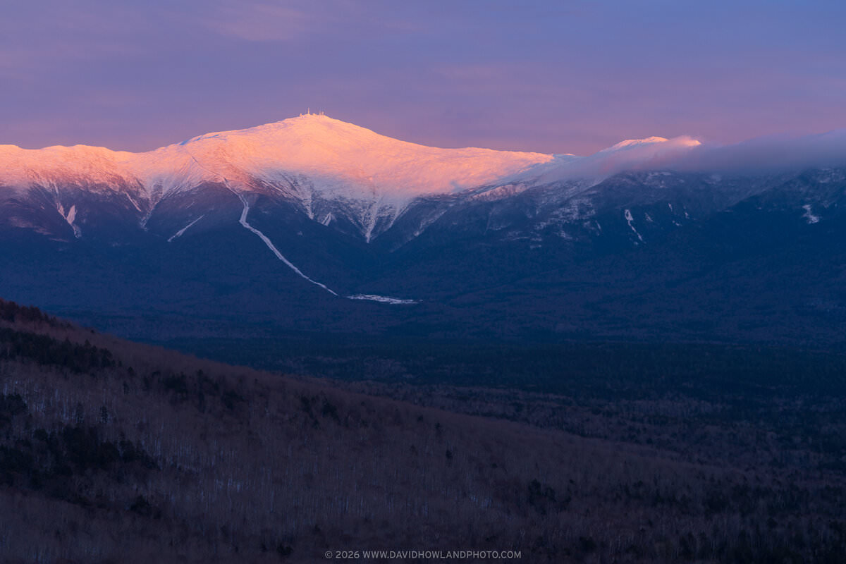 Mount Washington's snow-covered summit and surrounding peaks glow pink and orange from alpenglow at sunset, with communication towers visible on top and clouds drifting over Mount Monroe, while the foreground valleys remain in deep blue shadow.