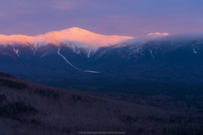 Mount Washington's snow-covered summit and surrounding peaks glow pink and orange from alpenglow at sunset, with communication towers visible on top and clouds drifting over Mount Monroe, while the foreground valleys remain in deep blue shadow.