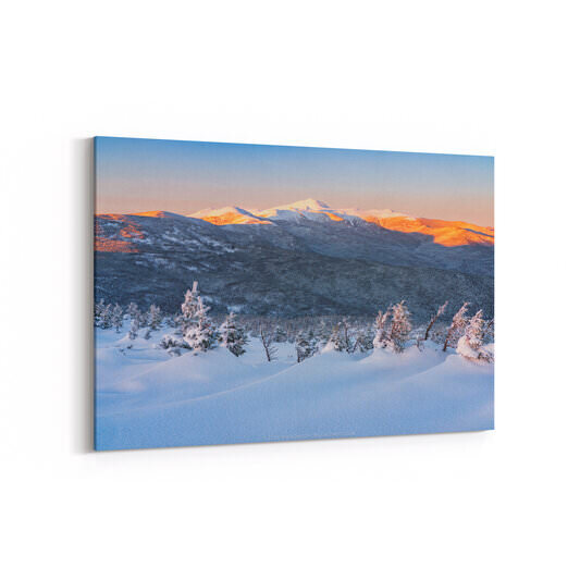 A winter landscape photograph of New Hampshire's Southern Presidential Range at sunset, featuring deep snow drifts and frost-covered evergreen trees in the foreground, layered snow-covered mountain ridges in the middle ground, and the high peaks bathed in warm alpenglow orange and pink light against a clear blue sky.