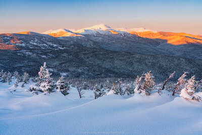 A winter landscape photograph of New Hampshire's Southern Presidential Range at sunset, featuring deep snow drifts and frost-covered evergreen trees in the foreground, layered snow-covered mountain ridges in the middle ground, and the high peaks bathed in warm alpenglow orange and pink light against a clear blue sky.