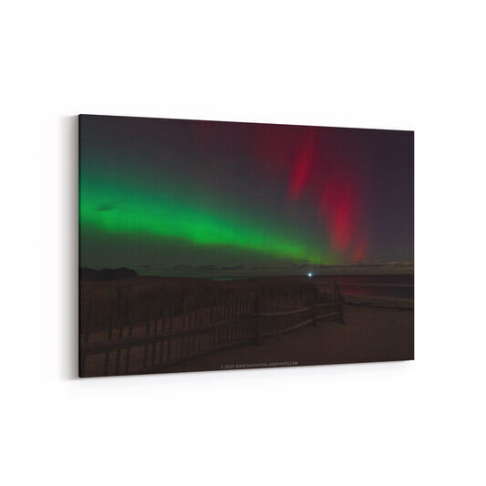The aurora borealis displays vibrant green and red curtains of light across a starry night sky above Coast Guard Beach, with a wooden fence and boardwalk silhouetted in the dark foreground and a distant light visible on the horizon.