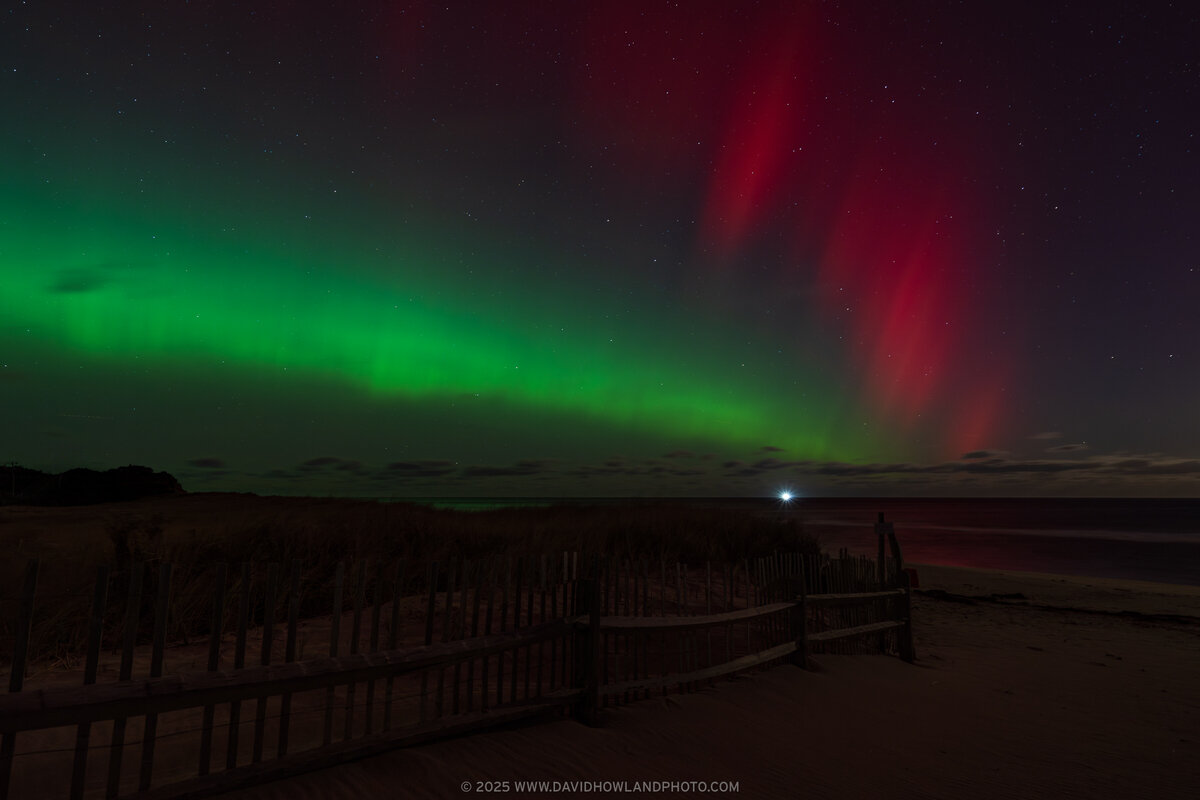 The aurora borealis displays vibrant green and red curtains of light across a starry night sky above Coast Guard Beach, with a wooden fence and boardwalk silhouetted in the dark foreground and a distant light visible on the horizon.