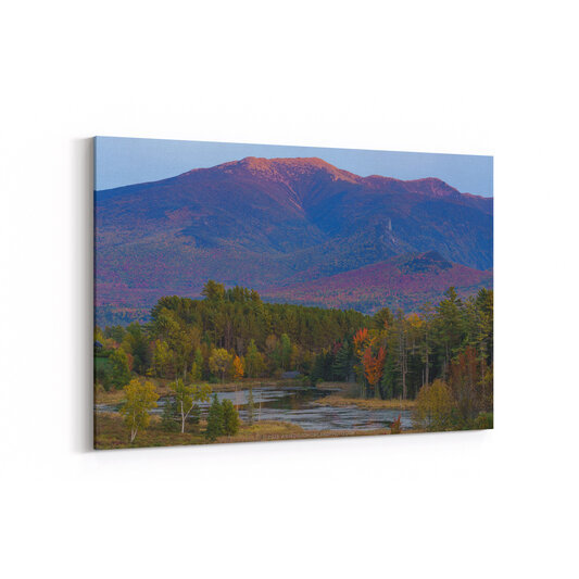 Mount Lafayette in New Hampshire's White Mountains rises majestically in the background with its ridgeline illuminated in warm sunset tones of pink and orange, while the foreground features a calm wetland area bordered by autumn foliage displaying vibrant reds, oranges, and yellows mixed with evergreen trees.
