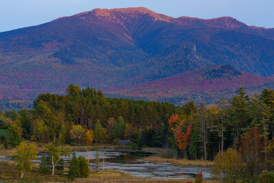 Mount Lafayette in New Hampshire's White Mountains rises majestically in the background with its ridgeline illuminated in warm sunset tones of pink and orange, while the foreground features a calm wetland area bordered by autumn foliage displaying vibrant reds, oranges, and yellows mixed with evergreen trees.