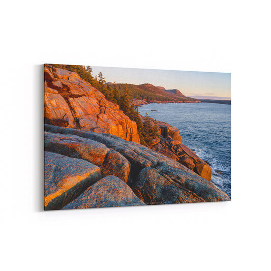 A sunrise view from Ocean Drive in Acadia National Park shows golden-orange granite cliffs on the left, calm blue ocean waters, and forested mountains across the bay under a soft pastel sky.