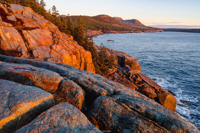A sunrise view from Ocean Drive in Acadia National Park shows golden-orange granite cliffs on the left, calm blue ocean waters, and forested mountains across the bay under a soft pastel sky.