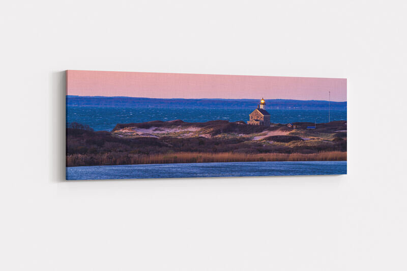 Block Island's North Lighthouse with its illuminated beacon sits on a coastal headland with dunes and golden grasses, framed by deep blue ocean waters and a soft pink sunset sky
