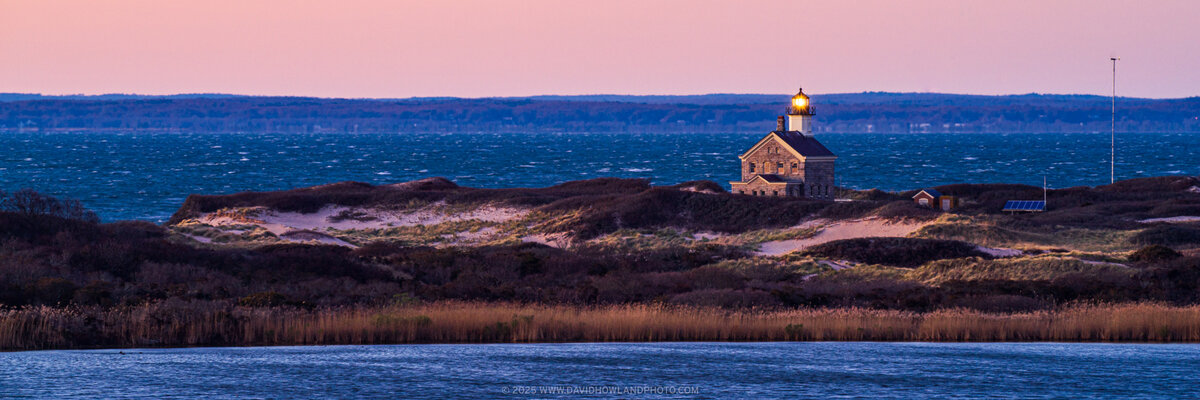 Block Island's North Lighthouse with its illuminated beacon sits on a coastal headland with dunes and golden grasses, framed by deep blue ocean waters and a soft pink sunset sky