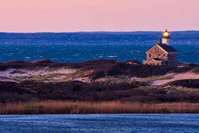Block Island's North Lighthouse with its illuminated beacon sits on a coastal headland with dunes and golden grasses, framed by deep blue ocean waters and a soft pink sunset sky